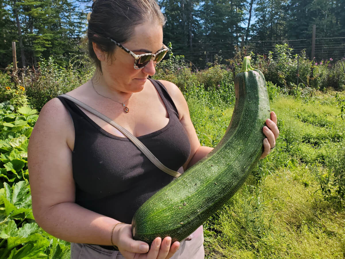 The image shows a woman standing outdoors holding a large, green, curving object that appears to be a fresh-picked cucumber or squash. The woman is wearing glasses and has a relaxed posture, possibly indicating she is at ease in this environment. She is surrounded by greenery, suggesting she might be in a garden or farm setting with a variety of plants growing around her. The focus on the large object held by the woman indicates it might be significant to the context of the image, perhaps as a result of gardening efforts or as part of a celebration of produce from a home-grown garden.